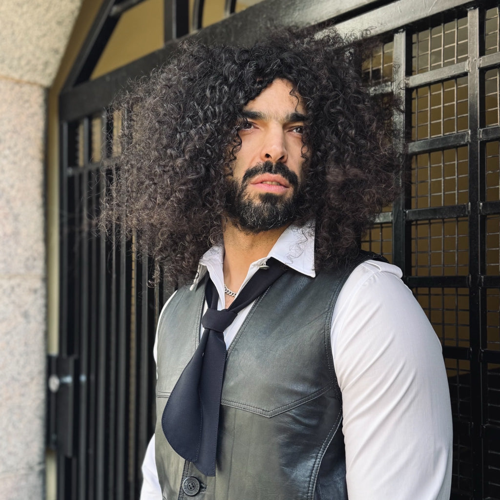 Man with curly hair and beard wearing a leather vest and tie in front of a building entrance.