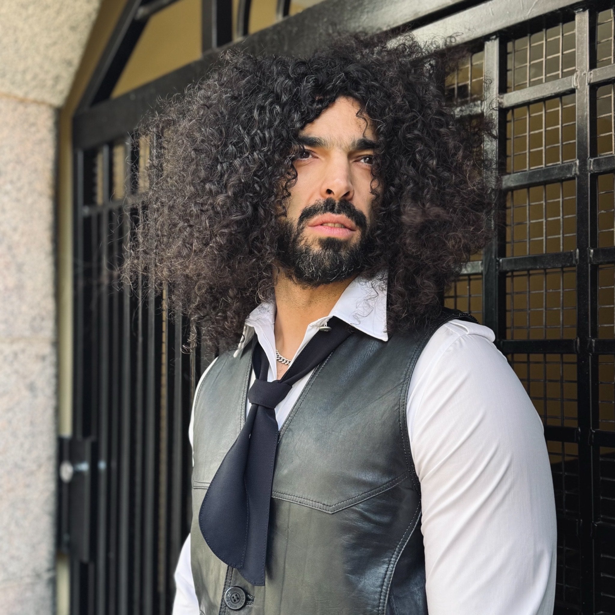 Man with curly hair and beard wearing a leather vest and tie in front of a building entrance.