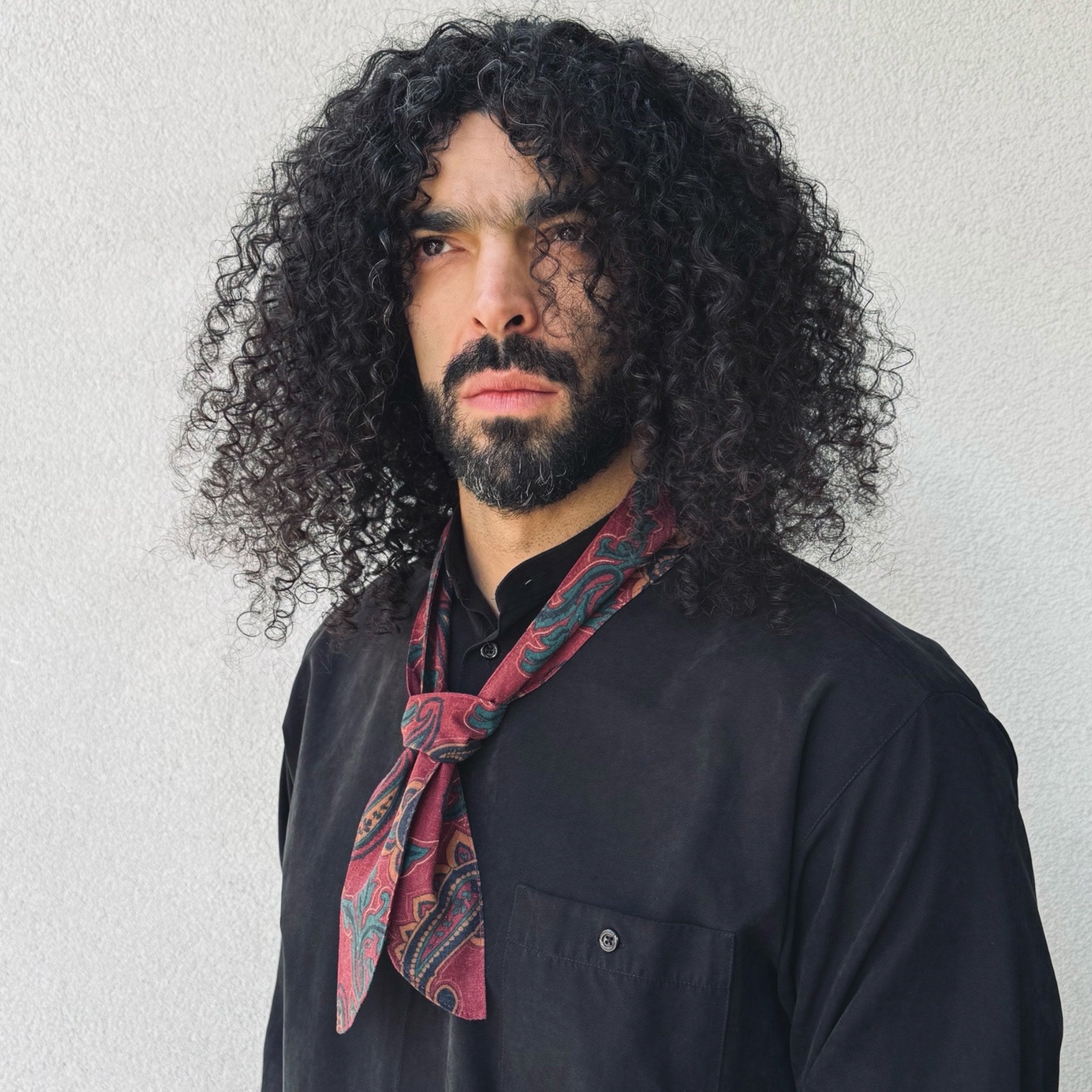 Man with curly hair and a beard wearing a dark shirt and patterned scarf against a light background