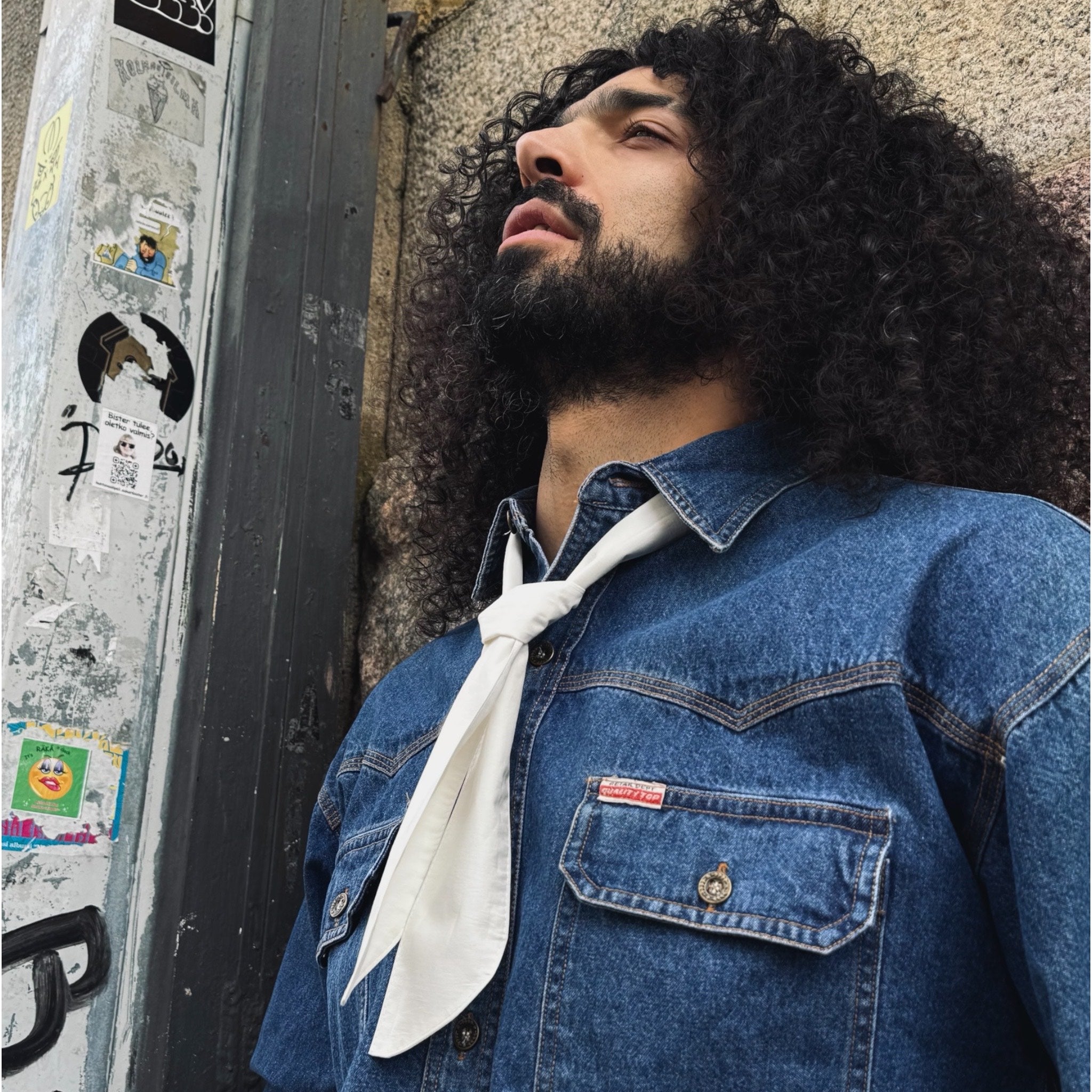 Man wearing a denim jacket with a white bandana, leaning against a textured wall.