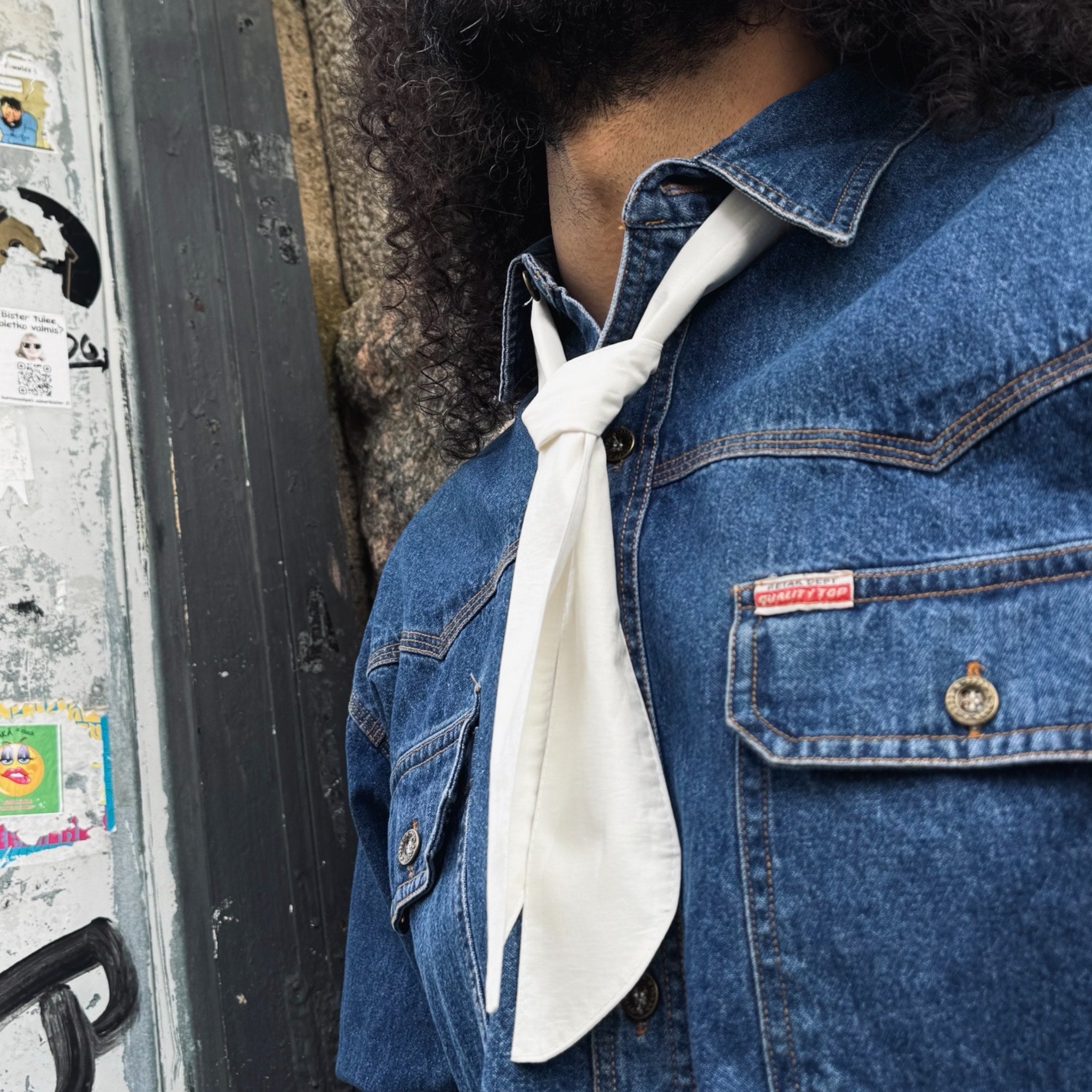 Person wearing a blue denim jacket with a white scarf, standing against a wall with posters.